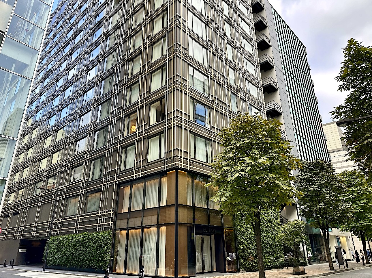 東京エディション銀座の外観。銀座の街角に佇む、黒を基調としたモダンな縦格子（ルーバー）デザインの建築。1階のエントランスからは温かみのある光が漏れ、着物で訪れる人々を優雅に迎え入れます。足元には植栽が配されています。The exterior of The Tokyo EDITION, Ginza, hotel. It features a modern architectural design with striking black vertical louvers integrated into the Ginza cityscape. The ground floor entrance glows with warm light, elegantly welcoming guests, including those visiting in kimono.