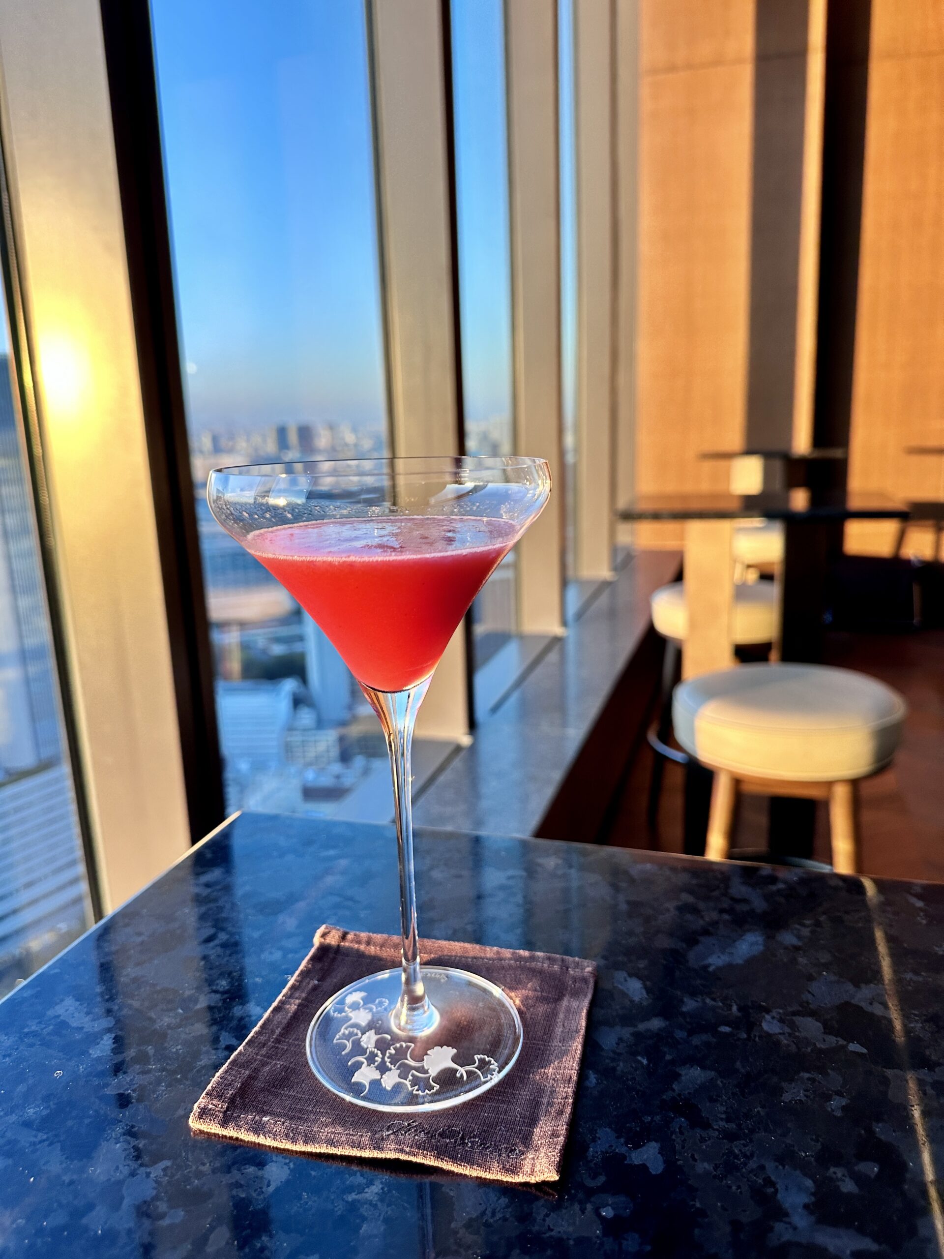 The Okura Tokyo 41階「バーラウンジ スターライト」の窓辺に置かれた、夕景に映える鮮やかな赤いカクテル A vivid red cocktail by the window at Bar Lounge Starlight on the 41st floor of The Okura Tokyo, illuminated by the sunset.