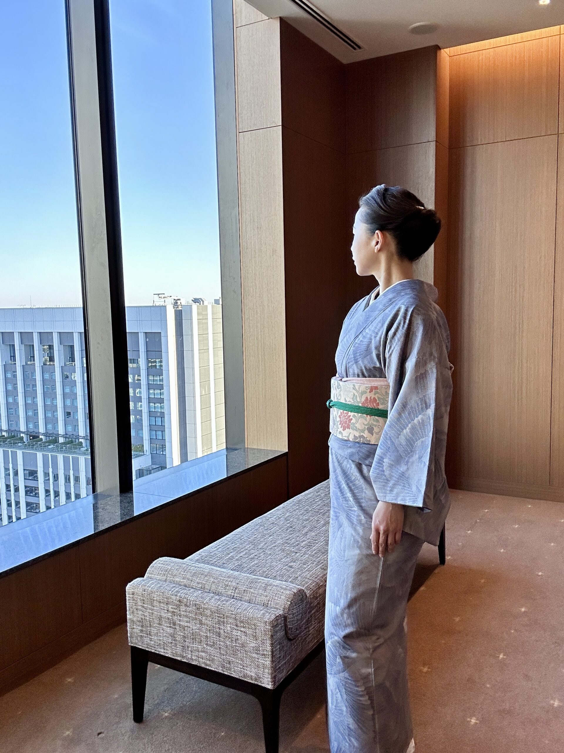 The Okura Tokyoの窓辺で、薄紫の都喜ヱ門の大島紬と吉祥文の名古屋帯を纏い、東京の景色を眺める女性 A woman wearing a pale purple Tokiemon Oshima tsumugi kimono with a Nagoya obi featuring auspicious motifs, looking out over Tokyo at The Okura Tokyo